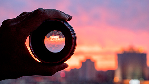 A hand holding a camera lens with a sunsetting urban background.