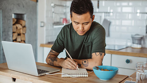 A young man sitting at a kitchen counter with an open laptop and writing in a notebook