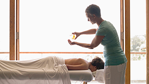 Massage therapist pouring massage oil in her hand while a client lies on a massage table.