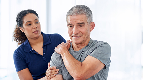An older man holding his shoulder while a physical therapist assists him.