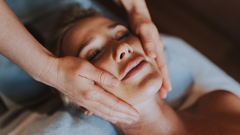 A woman lays supine while her massage therapist performs a facial massage.