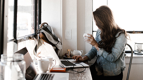 A woman sits at her home office surrounded by tech equipment while drinking coffee.
