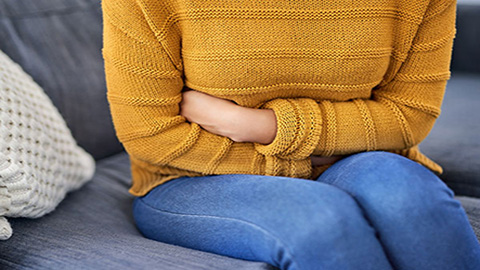 A woman in a yellow-knit sweater sits on a coach with her arms crossed over her abdomen.