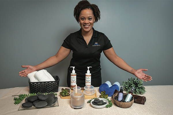 A Black female Massage Heights employee displaying various massage oils, stones, and towels.