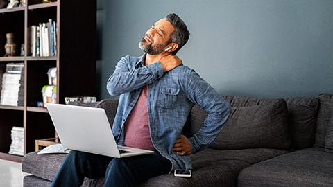 A man sitting on the couch working on his laptop grabbing his left shoulder in pain.