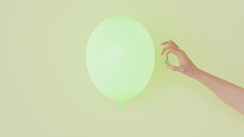 A hand holds a pin ready to pop a balloon set against a yellow background.