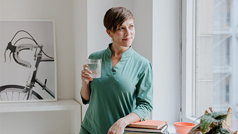 A woman stands looking out the window while holding a glass of water.
