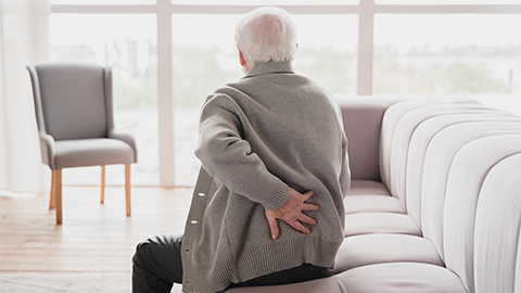 An elderly man sitting on the couch holding his lower back in pain.