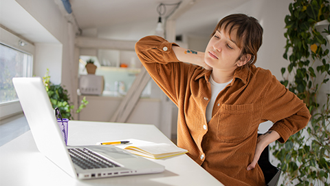 A woman sitting at a desk working on her computer, grabbing her shoulders and lower back in pain.