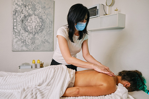 Massage therapist wearing a face mask while massaging a client's back in a treatment room.