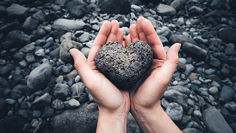 Hands holding a heart-shaped rock above a river. 