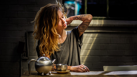 Woman with tattoos sitting at a table rubbing her shoulder.