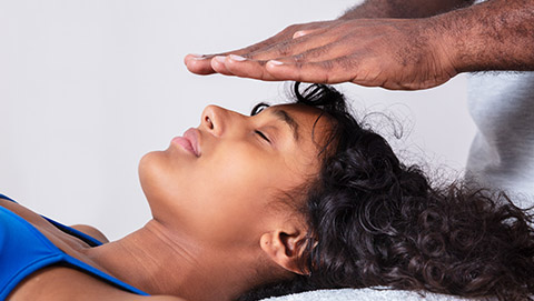 Black woman lying on her back while reiki practitioner performs reiki with hands above her head.
