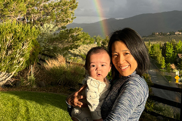 Kei Yumoto holds her baby while posing for a picture in front of a double rainbow that appears over a valley.
